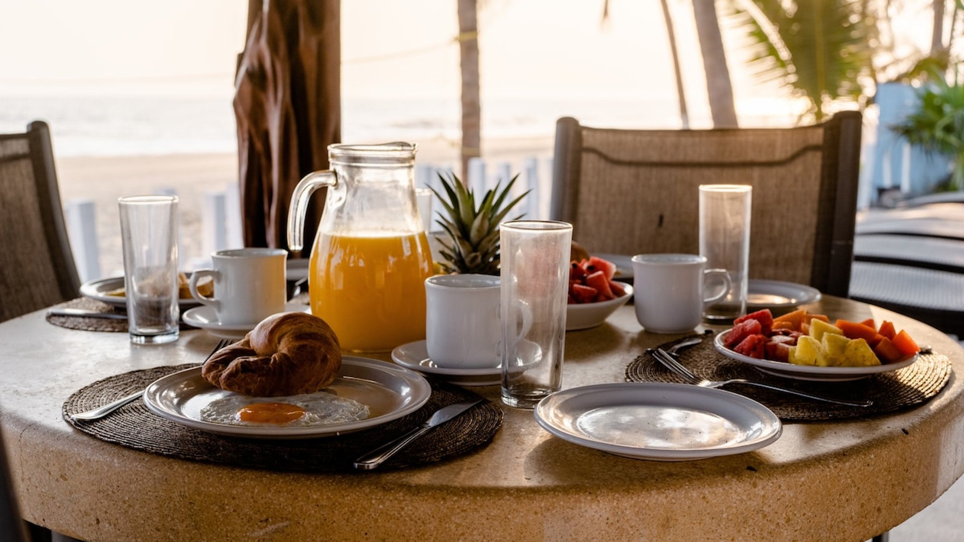 Mesa redonda de piedra con desayuno frente a la playa: jugo, fruta, croissant y huevos con palmeras al fondo