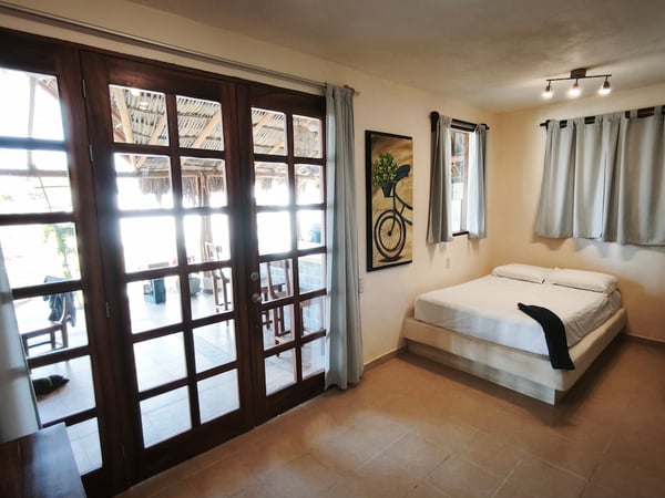 Recámara luminosa con cama doble en plataforma, puertas francesas de madera hacia terraza con palapa al fondo