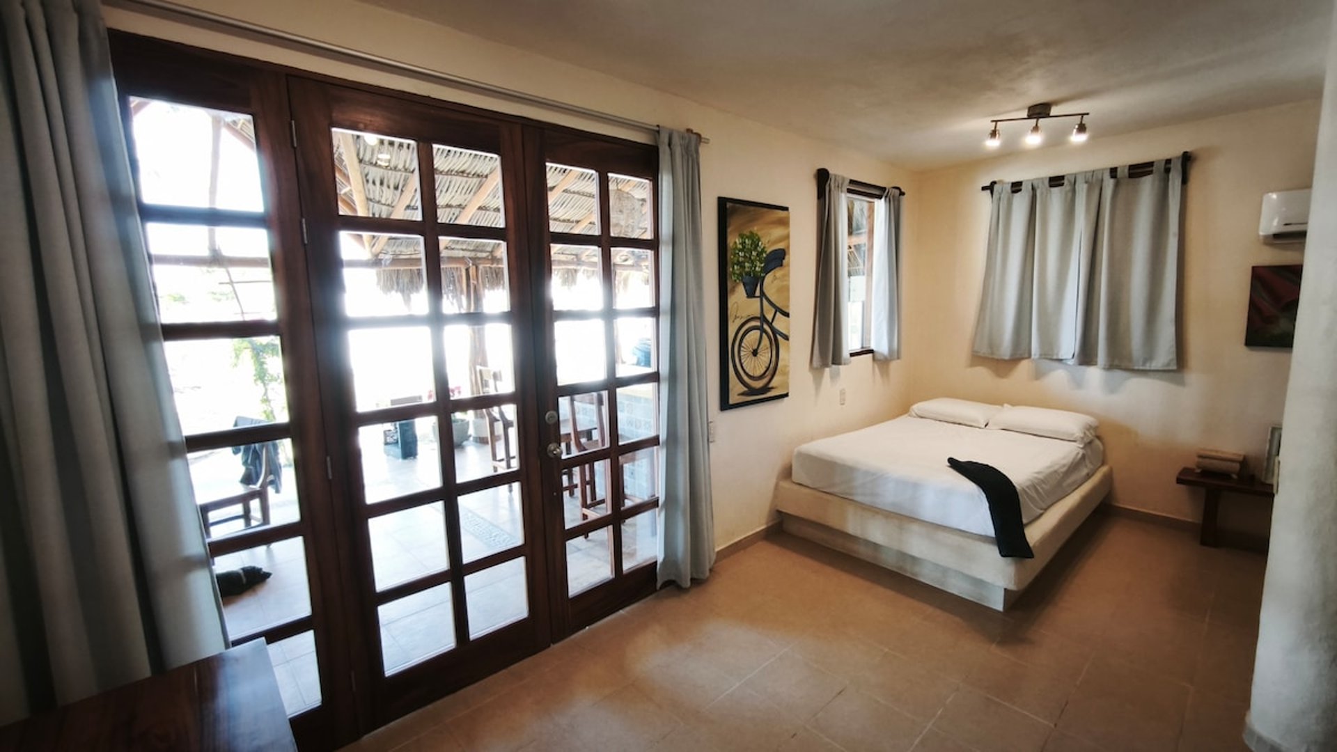Recámara luminosa con cama doble en plataforma, puertas francesas de madera hacia terraza con palapa al fondo