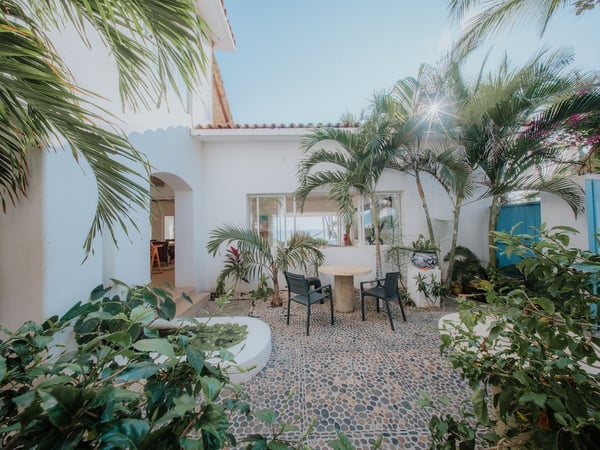 Patio interior empedrado con mesa y sillas rodeado de palmeras y vegetación tropical, villa blanca con vista al mar al fondo