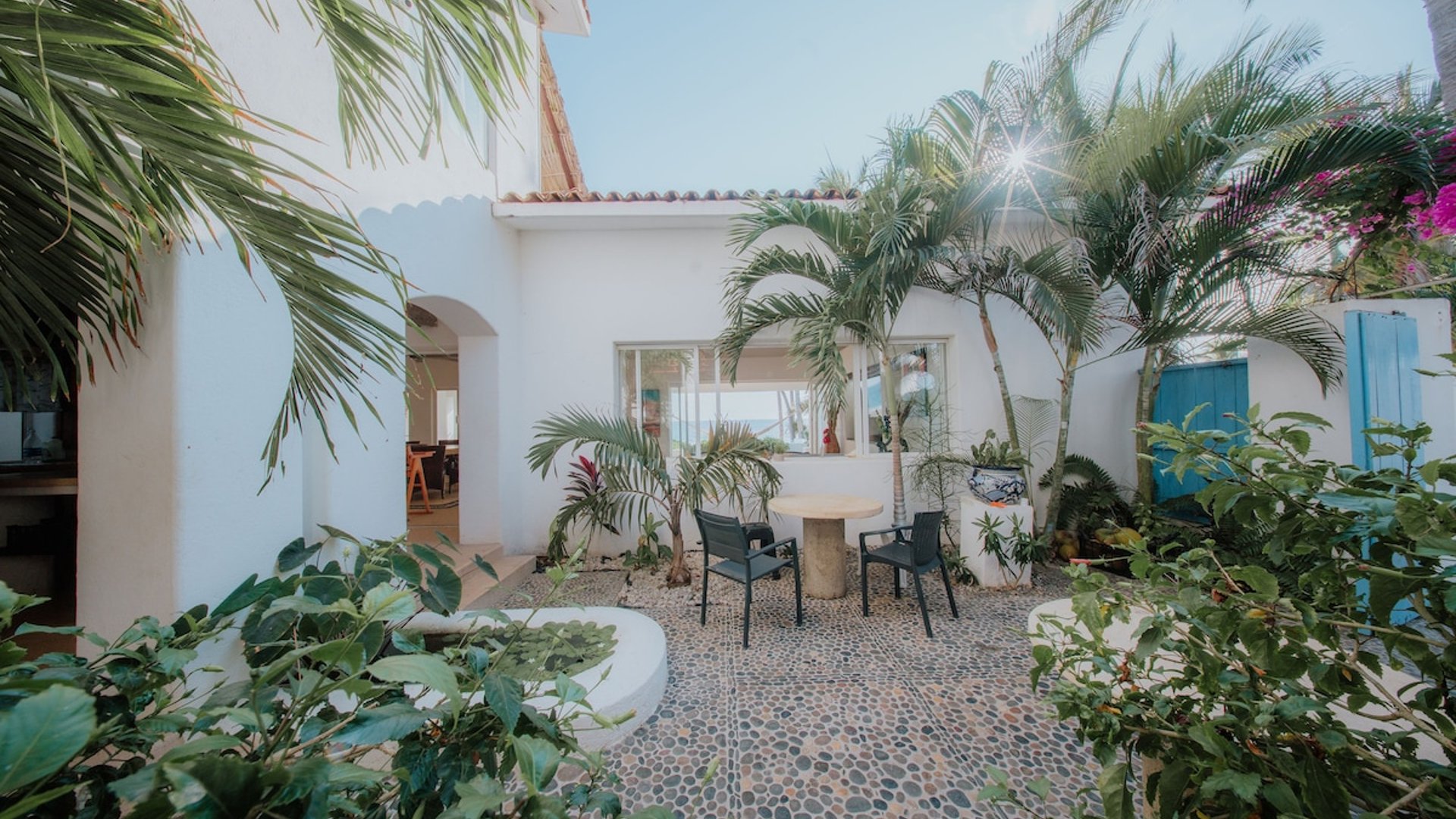 Patio interior empedrado con mesa y sillas rodeado de palmeras y vegetación tropical, villa blanca con vista al mar al fondo