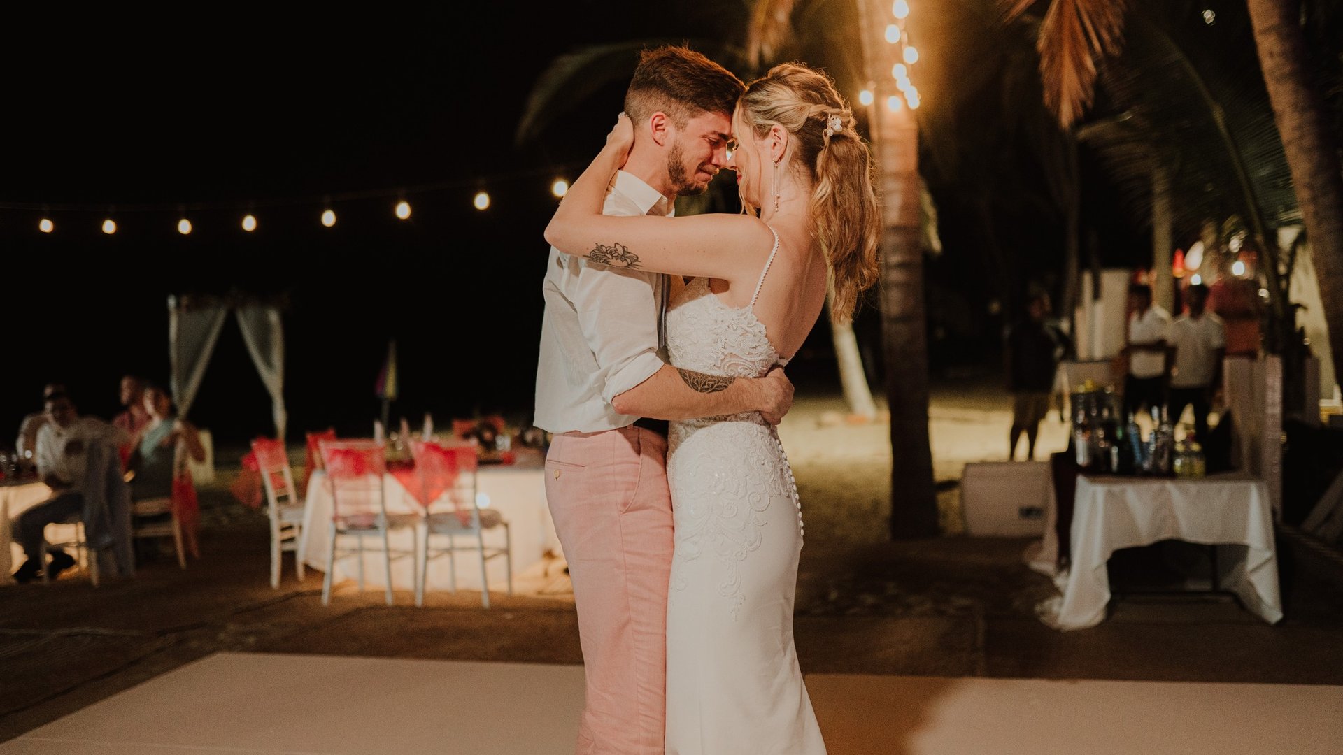 Primer baile de bodas en terraza nocturna con palmeras y luces colgantes frente a la playa