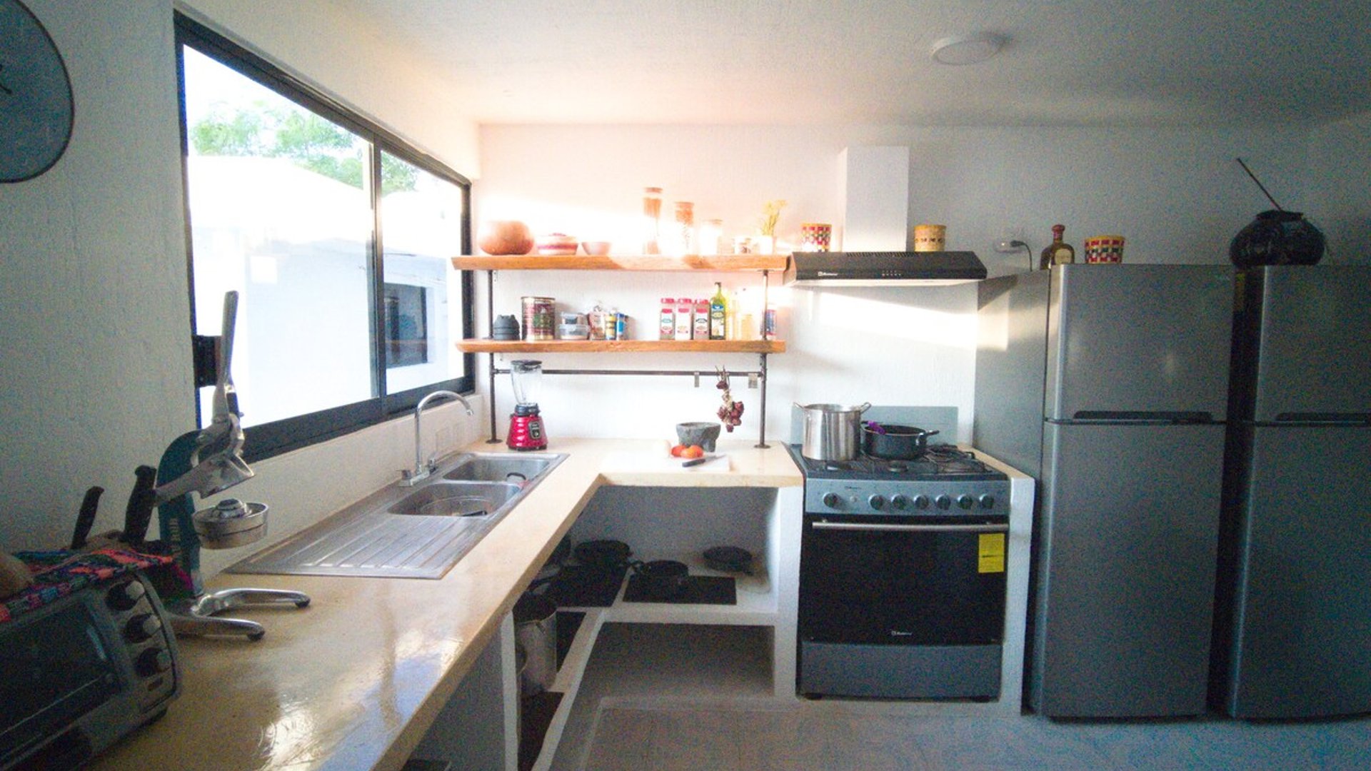 Cocina equipada en L con estufa, refrigerador de acero, repisas de madera y ventana con luz natural