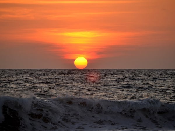 Sol redondo y encendido tocando el horizonte del Pacífico mientras una ola rompe en primer plano, cielo naranja intenso