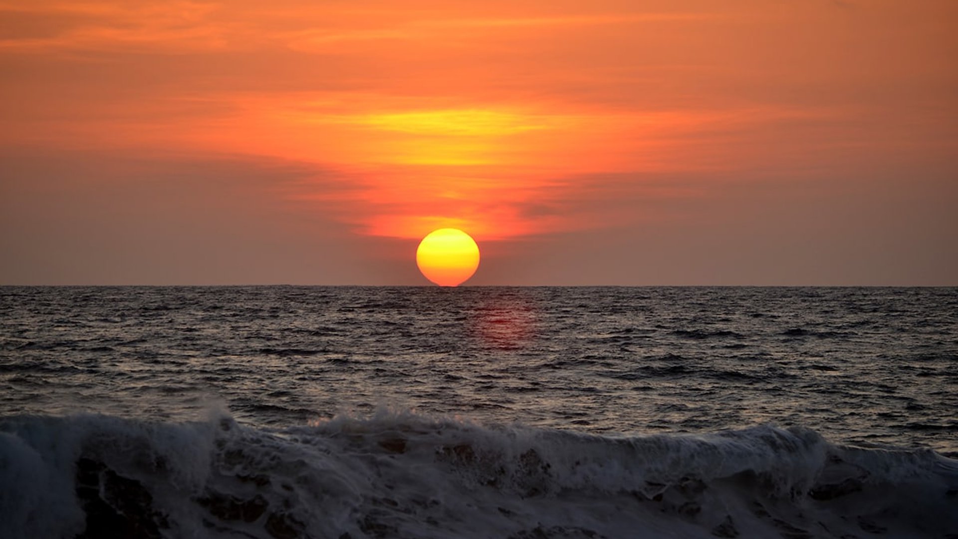 Sol redondo y encendido tocando el horizonte del Pacífico mientras una ola rompe en primer plano, cielo naranja intenso