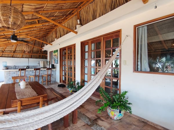 Palapa abierta con hamaca, mesa de madera rústica, cocina de barra y puertas francesas de madera hacia el interior