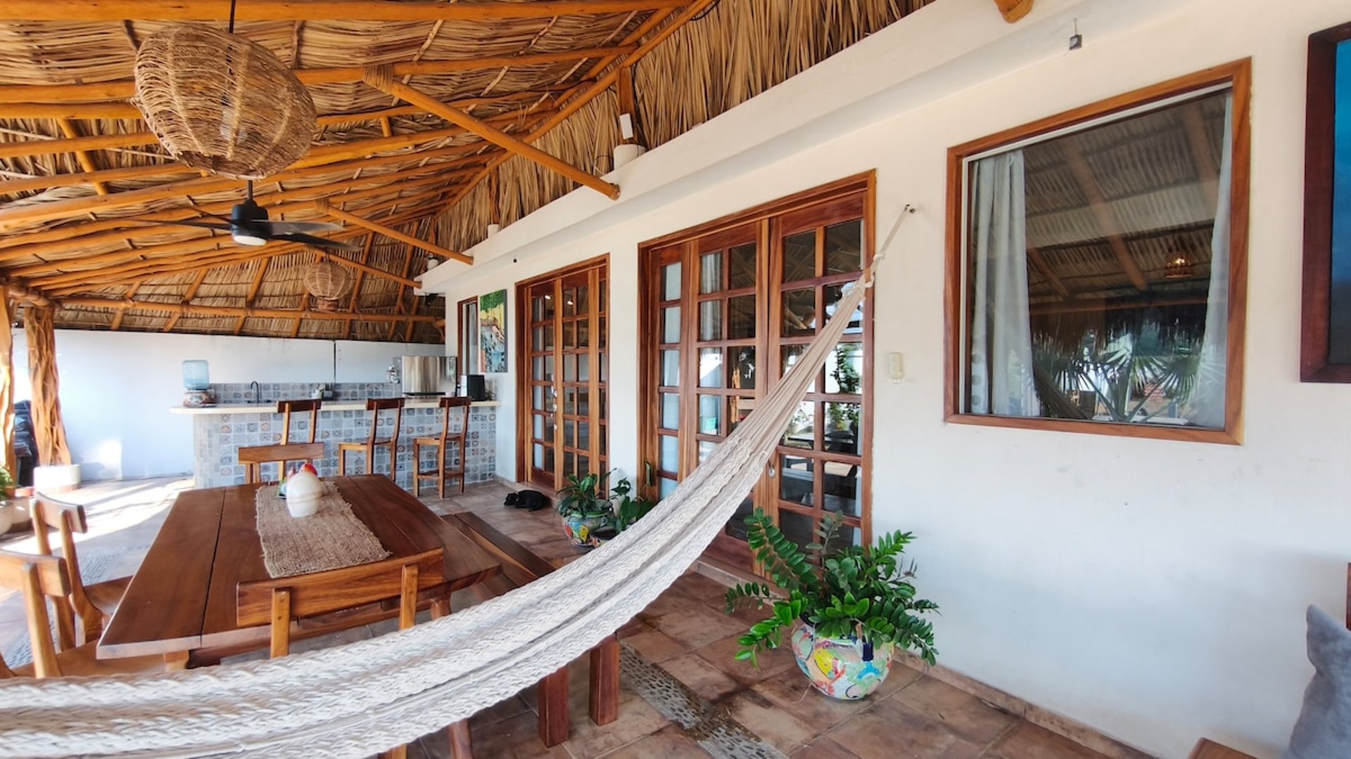 Palapa abierta con hamaca, mesa de madera rústica, cocina de barra y puertas francesas de madera hacia el interior