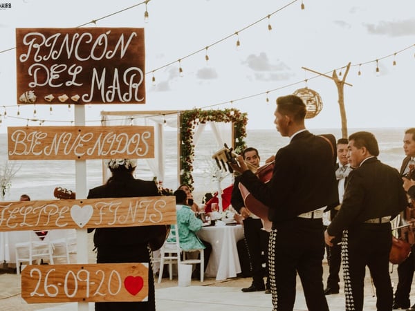 Boda en la playa con mariachi, arco floral y letreros de bienvenida frente al mar al atardecer