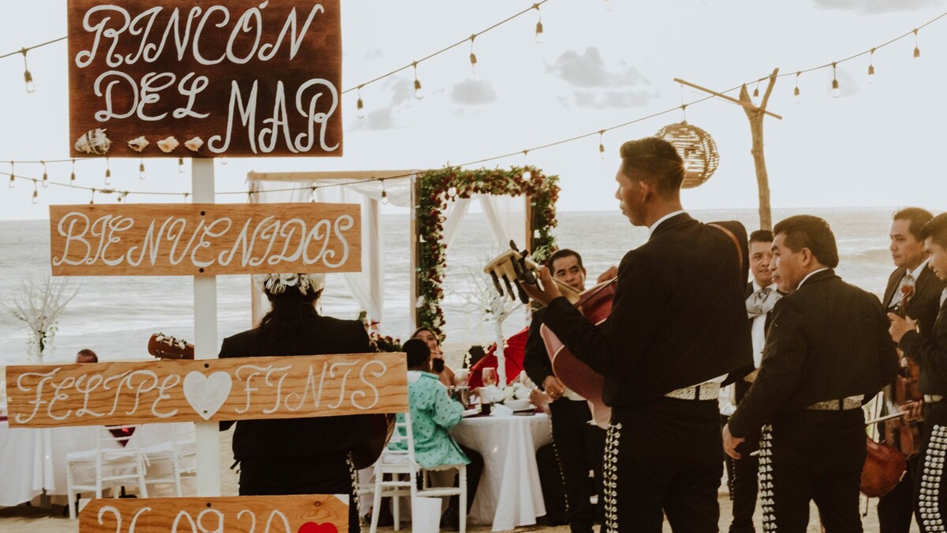 Boda en la playa con mariachi, arco floral y letreros de bienvenida frente al mar al atardecer