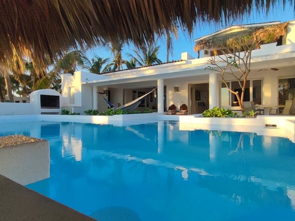 Alberca amplia frente a villa blanca con hamaca y palapa, palmeras al fondo en luz cálida de tarde