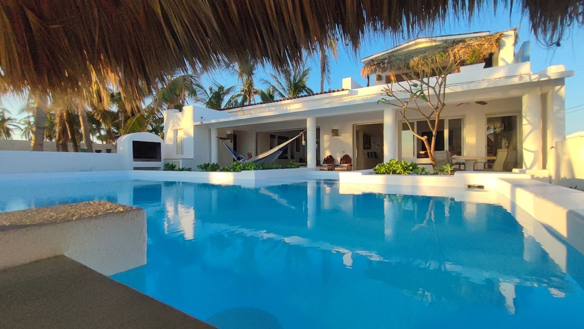Alberca amplia frente a villa blanca con hamaca y palapa, palmeras al fondo en luz cálida de tarde