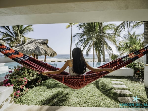 Hamaca artesanal bajo terraza con vista a alberca, palapa y mar al fondo entre palmeras tropicales