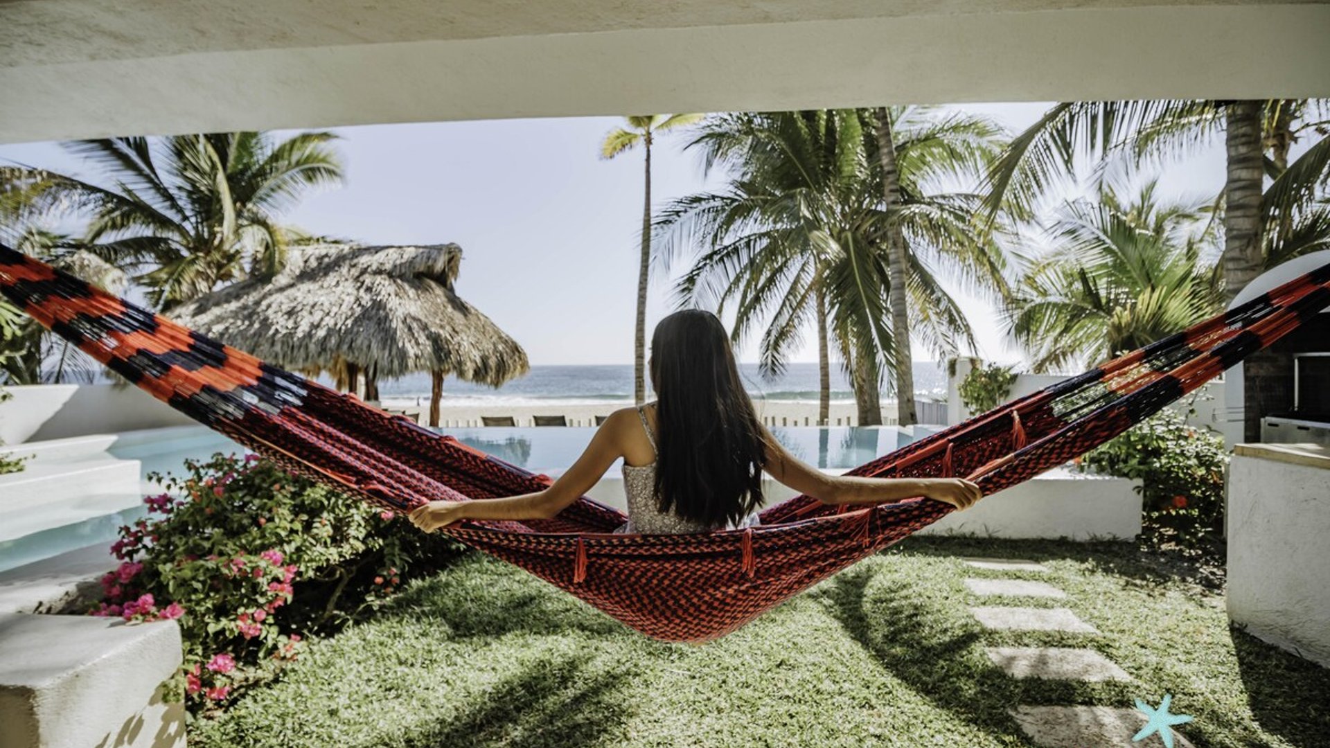 Hamaca artesanal bajo terraza con vista a alberca, palapa y mar al fondo entre palmeras tropicales