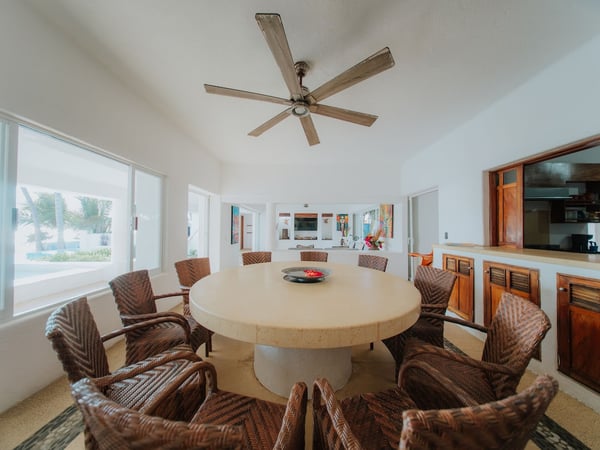 Comedor interior con mesa redonda de concreto y sillas de ratán para ocho personas, ventilador de techo y vista hacia jardín