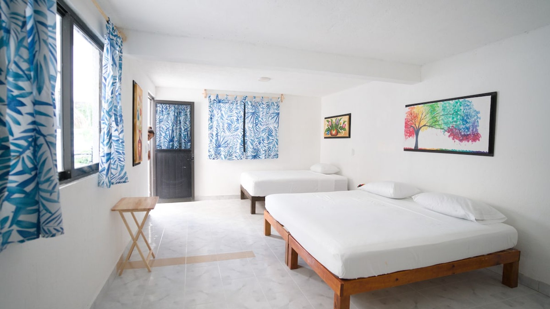 Recámara amplia y luminosa con dos camas individuales en base de madera, cortinas azules tropicales y cuadros coloridos en paredes blancas