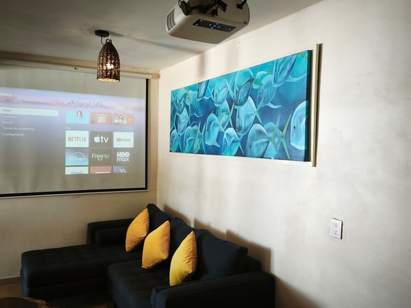 Sala de cine privada con proyector, pantalla Roku y sofá oscuro con cojines amarillos junto a cuadro marino