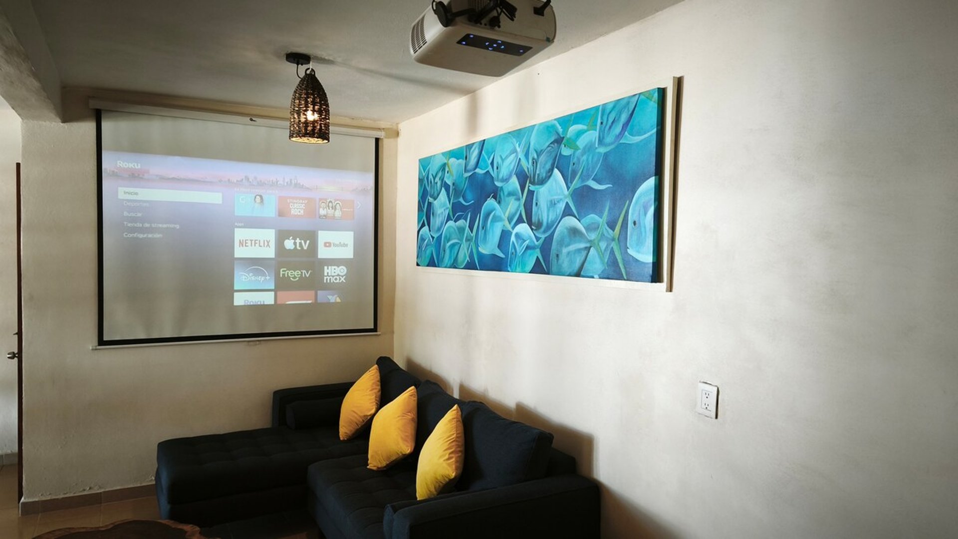 Sala de cine privada con proyector, pantalla Roku y sofá oscuro con cojines amarillos junto a cuadro marino