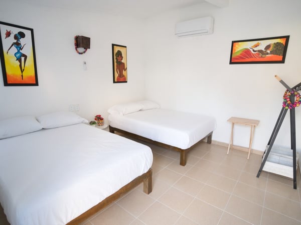 Recámara con dos camas individuales, ropa de cama blanca, arte afrolatino en paredes y aire acondicionado