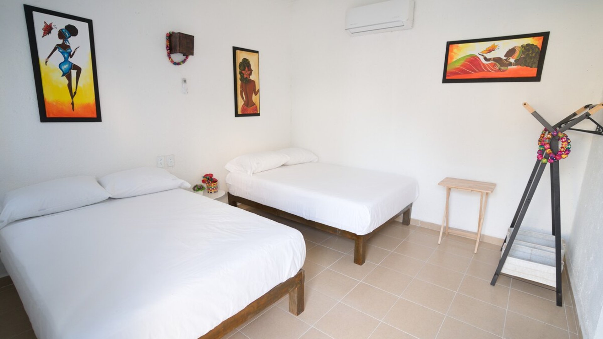 Recámara con dos camas individuales, ropa de cama blanca, arte afrolatino en paredes y aire acondicionado