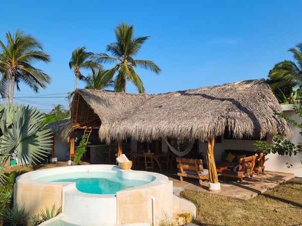 Jacuzzi redondo frente a palapa con techo de palma, hamaca y sala exterior rodeada de palmeras bajo cielo azul