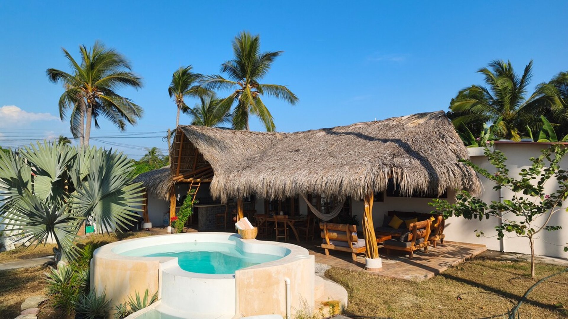 Jacuzzi redondo frente a palapa con techo de palma, hamaca y sala exterior rodeada de palmeras bajo cielo azul