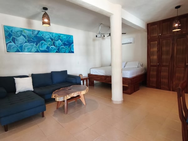 Estudio tipo loft con sofá azul, mesa de madera rústica, cama doble y armario de madera oscura; cuadro marino en pared blanca