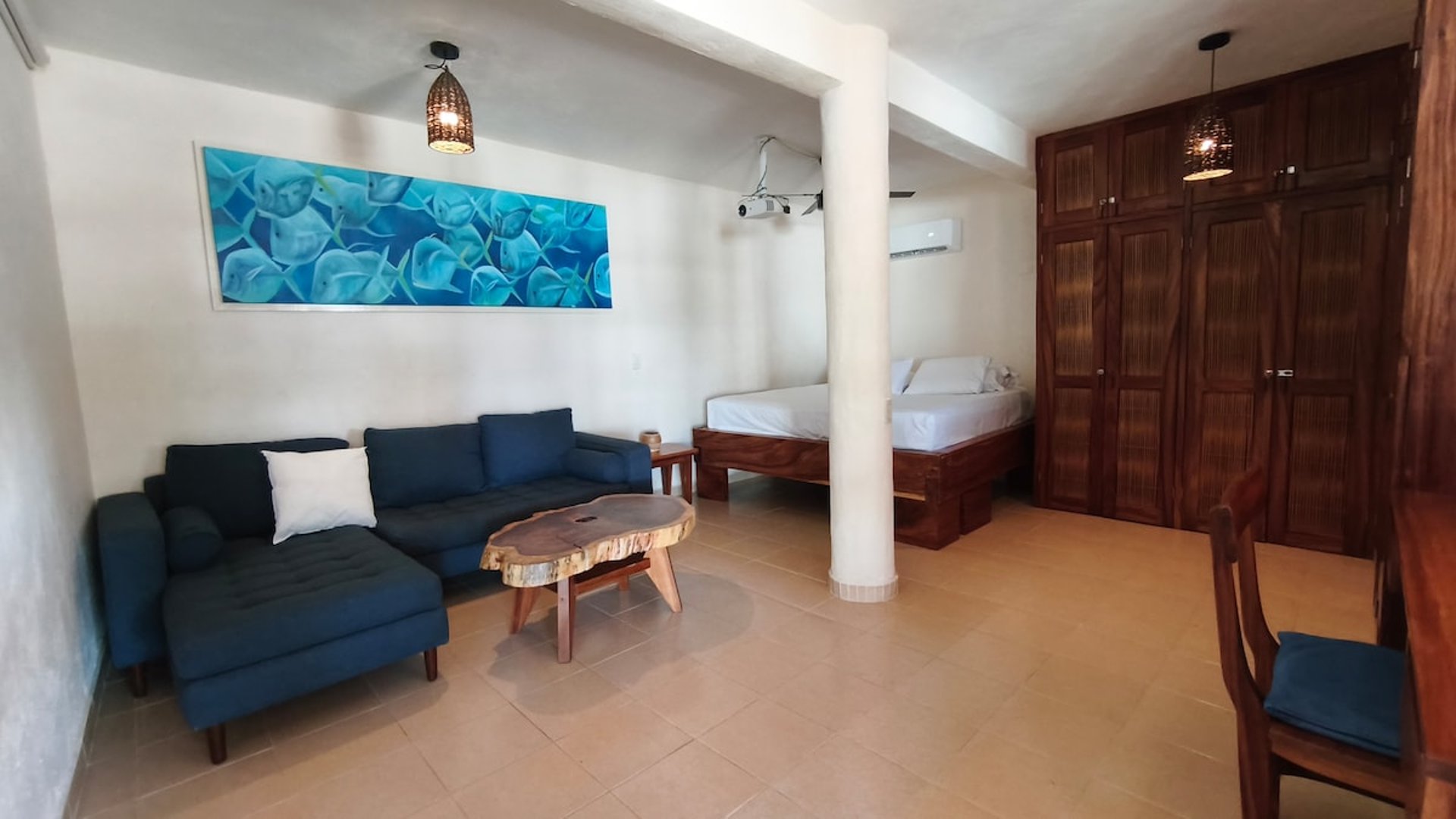 Estudio tipo loft con sofá azul, mesa de madera rústica, cama doble y armario de madera oscura; cuadro marino en pared blanca