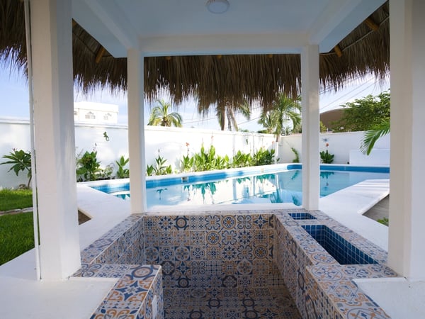 Jacuzzi con mosaicos talavera bajo palapa blanca y alberca al fondo rodeada de vegetación tropical