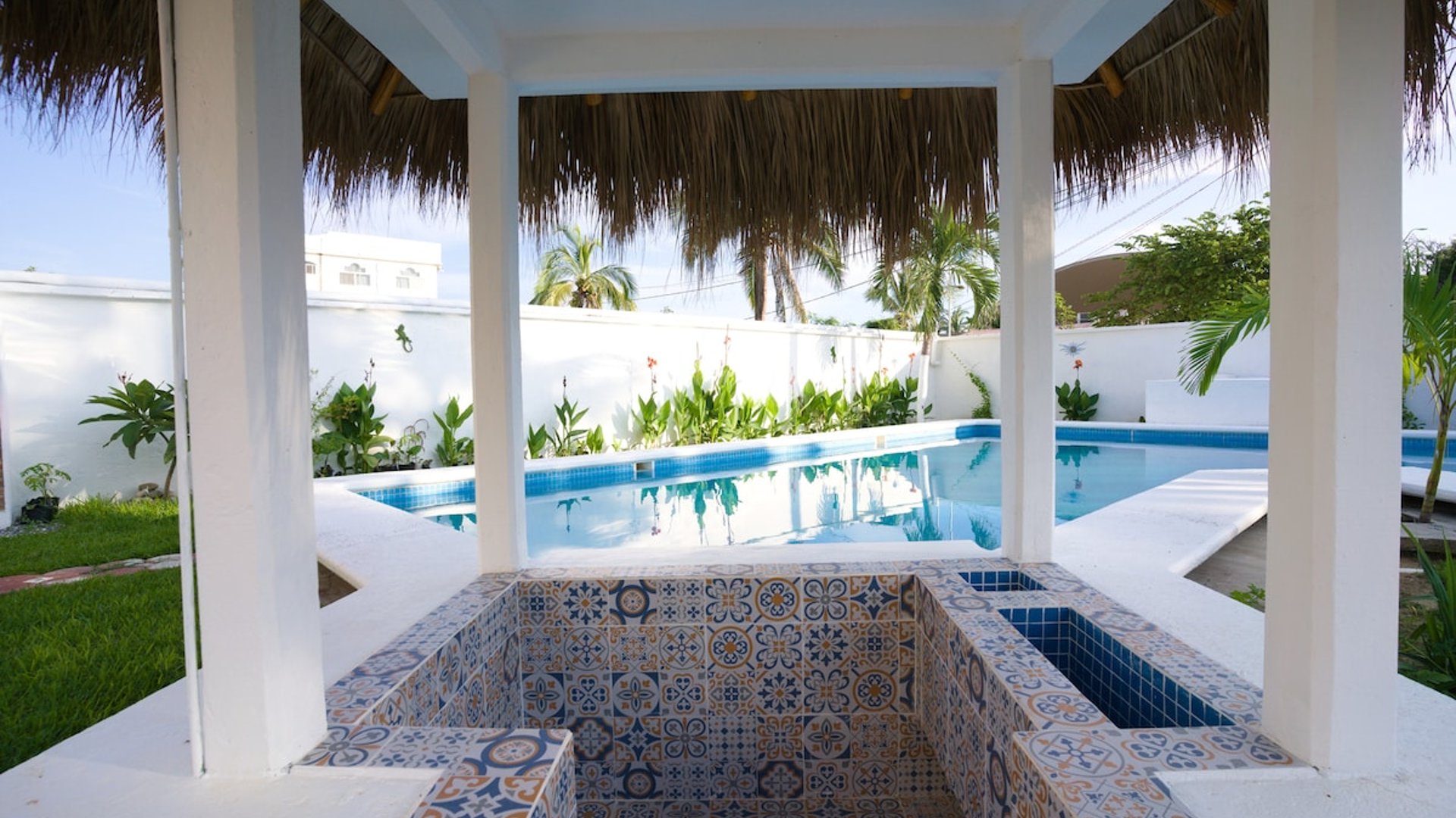 Jacuzzi con mosaicos talavera bajo palapa blanca y alberca al fondo rodeada de vegetación tropical