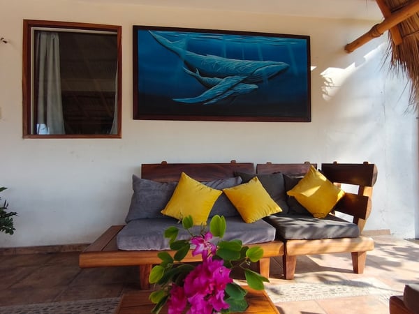 Sala exterior bajo palapa con sillones de madera, cojines amarillos y cuadro de ballenas azules en la pared