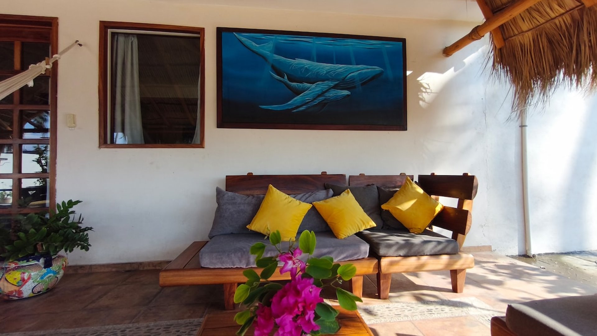 Sala exterior bajo palapa con sillones de madera, cojines amarillos y cuadro de ballenas azules en la pared