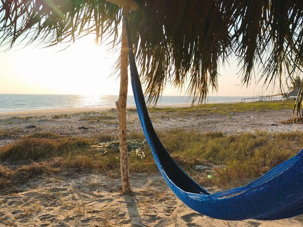 Hamaca azul bajo palapa frente al mar al atardecer, arena y horizonte dorado en la costa del Pacífico