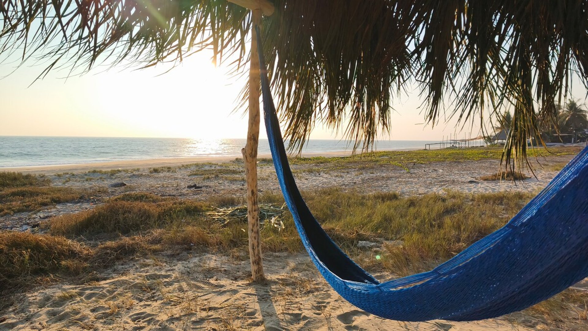 Hamaca azul bajo palapa frente al mar al atardecer, arena y horizonte dorado en la costa del Pacífico
