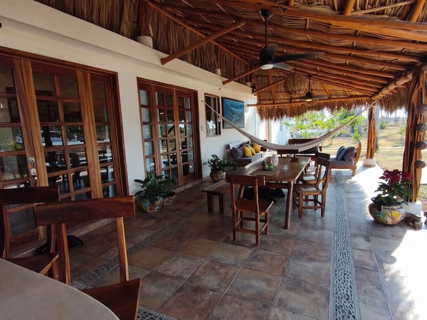 Terraza bajo palapa con comedor de madera, hamaca, sala exterior y ventiladores de techo, abierta al jardín