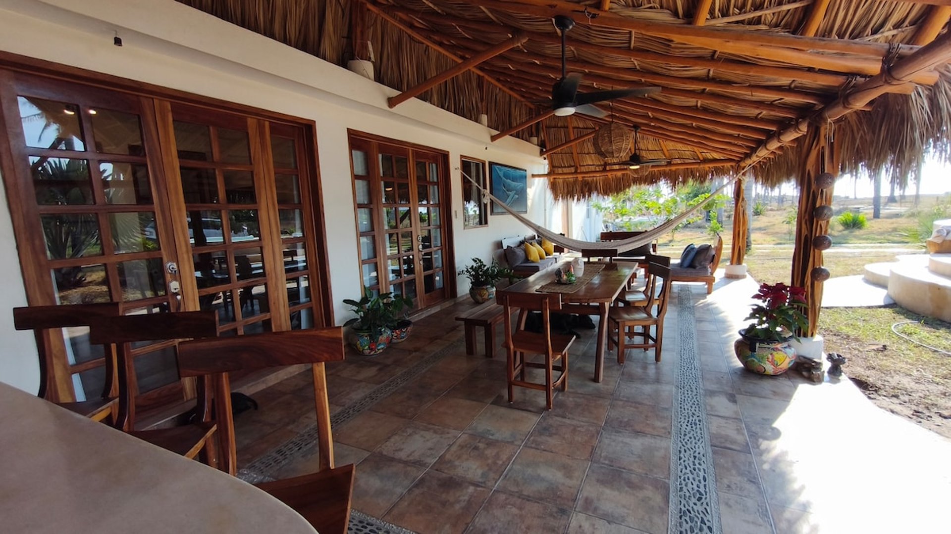 Terraza bajo palapa con comedor de madera, hamaca, sala exterior y ventiladores de techo, abierta al jardín
