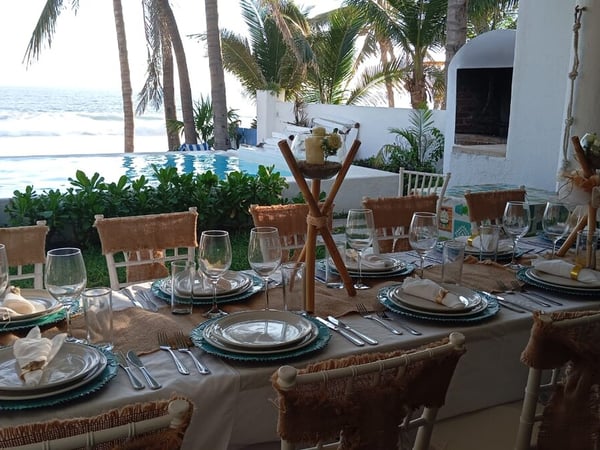 Mesa de banquete montada con vajilla elegante y centros de mesa frente a alberca infinity y mar con palmeras