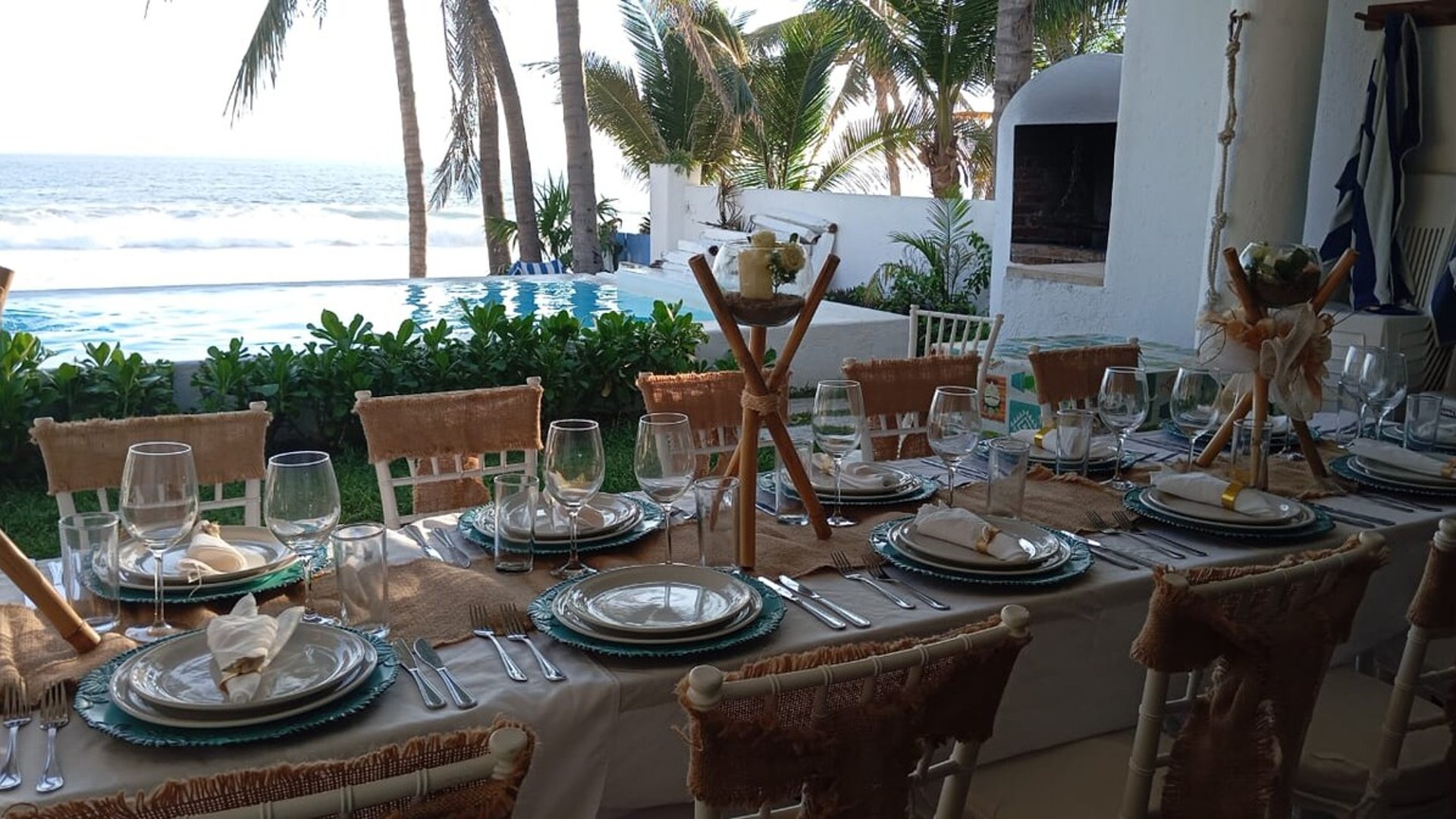 Mesa de banquete montada con vajilla elegante y centros de mesa frente a alberca infinity y mar con palmeras