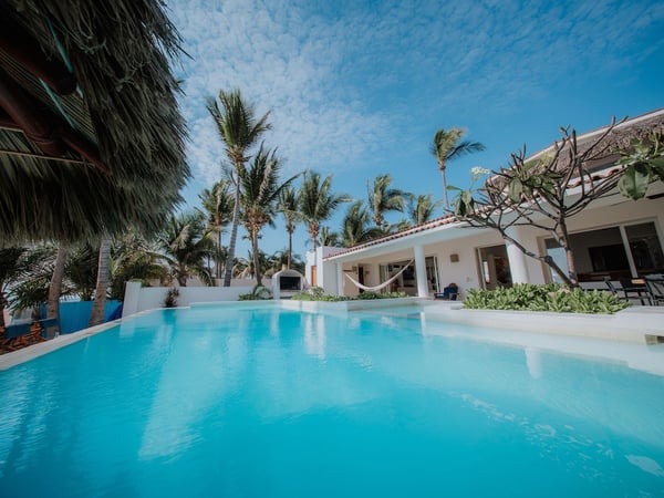 Alberca de aguas turquesas frente a villa blanca con hamaca, palmeras y cielo azul despejado