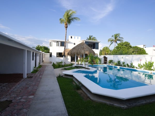 Vista del jardín interior con alberca azul, jacuzzi, palapa y edificio principal blanco de dos pisos bajo cielo despejado