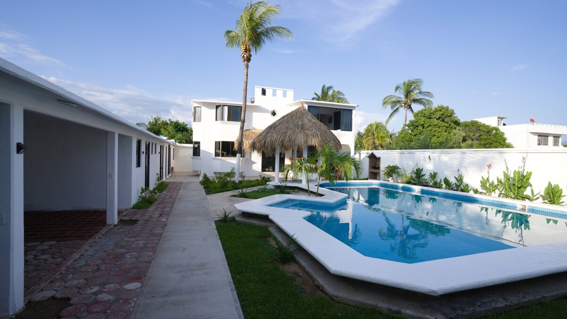 Vista del jardín interior con alberca azul, jacuzzi, palapa y edificio principal blanco de dos pisos bajo cielo despejado