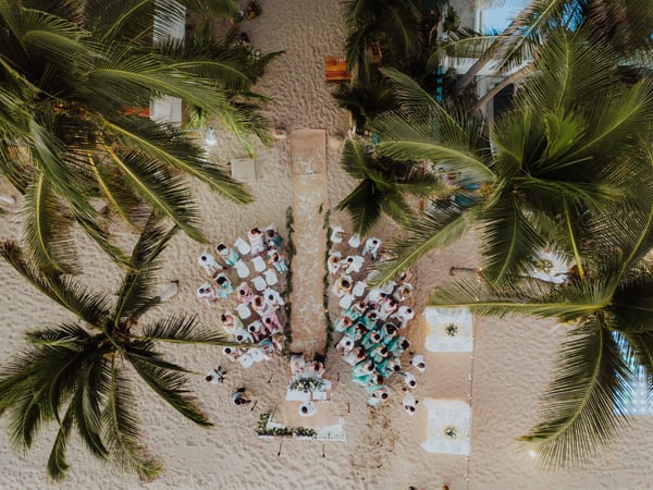 Vista aérea de ceremonia de boda en playa con pasillo de arena entre palmeras e invitados sentados