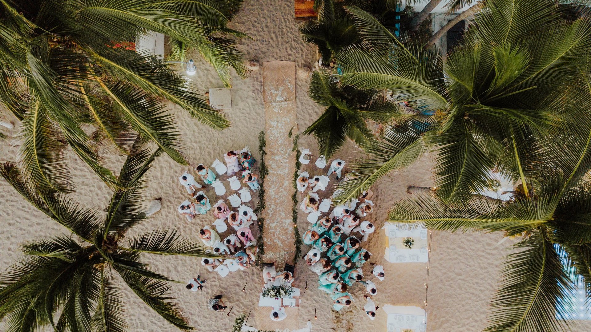 Vista aérea de ceremonia de boda en playa con pasillo de arena entre palmeras e invitados sentados
