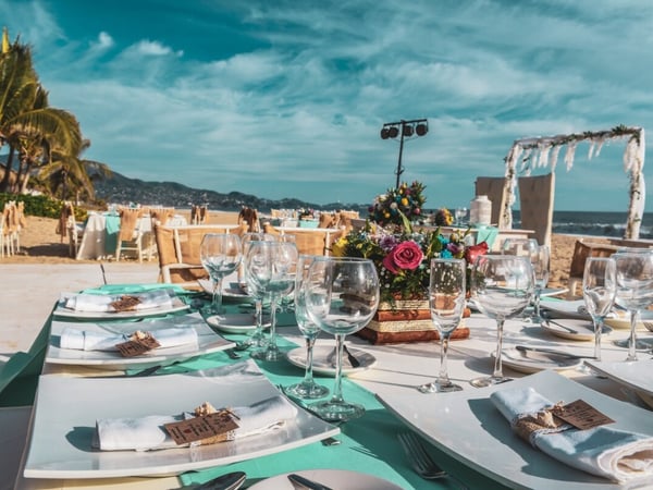 Montaje de boda en la playa con mesas decoradas en turquesa, copas de cristal, flores coloridas y arco nupcial frente al mar