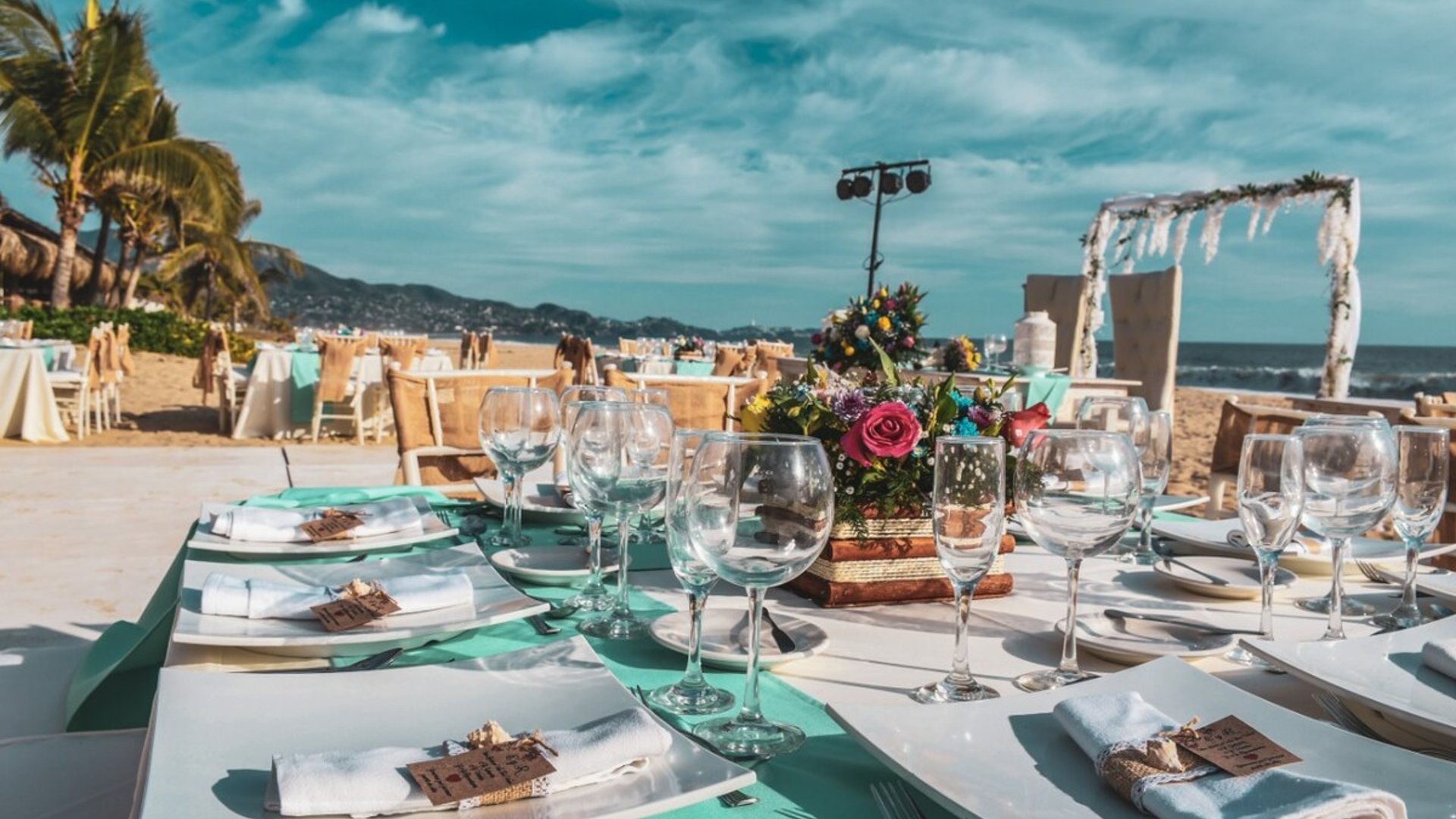 Montaje de boda en la playa con mesas decoradas en turquesa, copas de cristal, flores coloridas y arco nupcial frente al mar