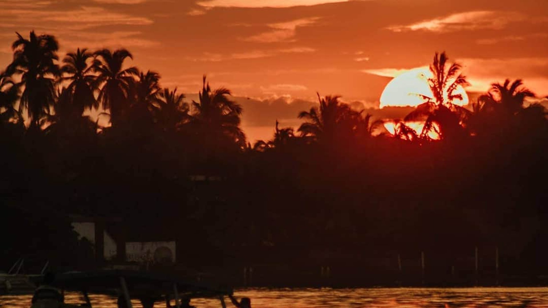 Silueta de lancha navegando en laguna al atardecer con palmeras y cielo rojo intenso