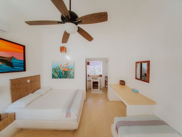 Recámara luminosa con cama doble, cabecera de mimbre, arte tropical en paredes blancas y ventilador de techo