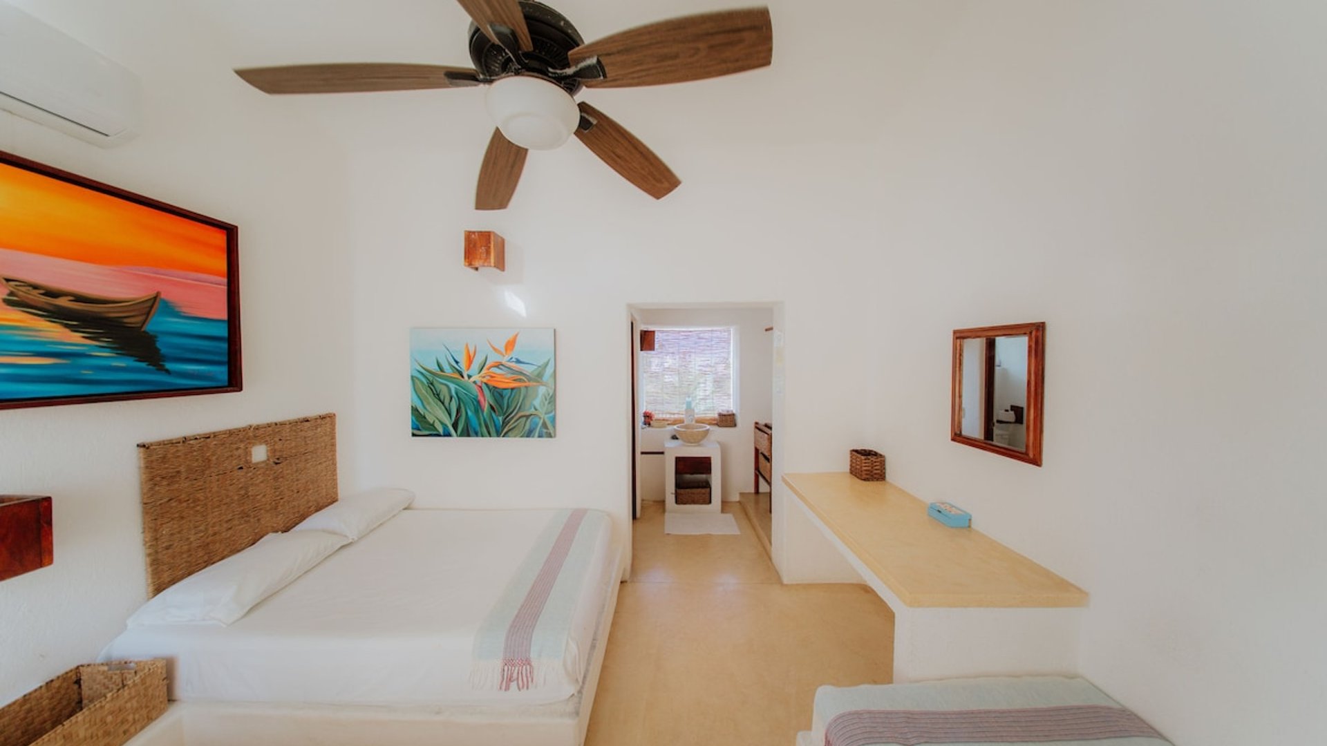 Recámara luminosa con cama doble, cabecera de mimbre, arte tropical en paredes blancas y ventilador de techo
