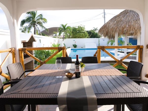 Comedor exterior techado con mesa de seis lugares, botella de vino y copa; al fondo alberca, palapa y palmeras en jardín soleado