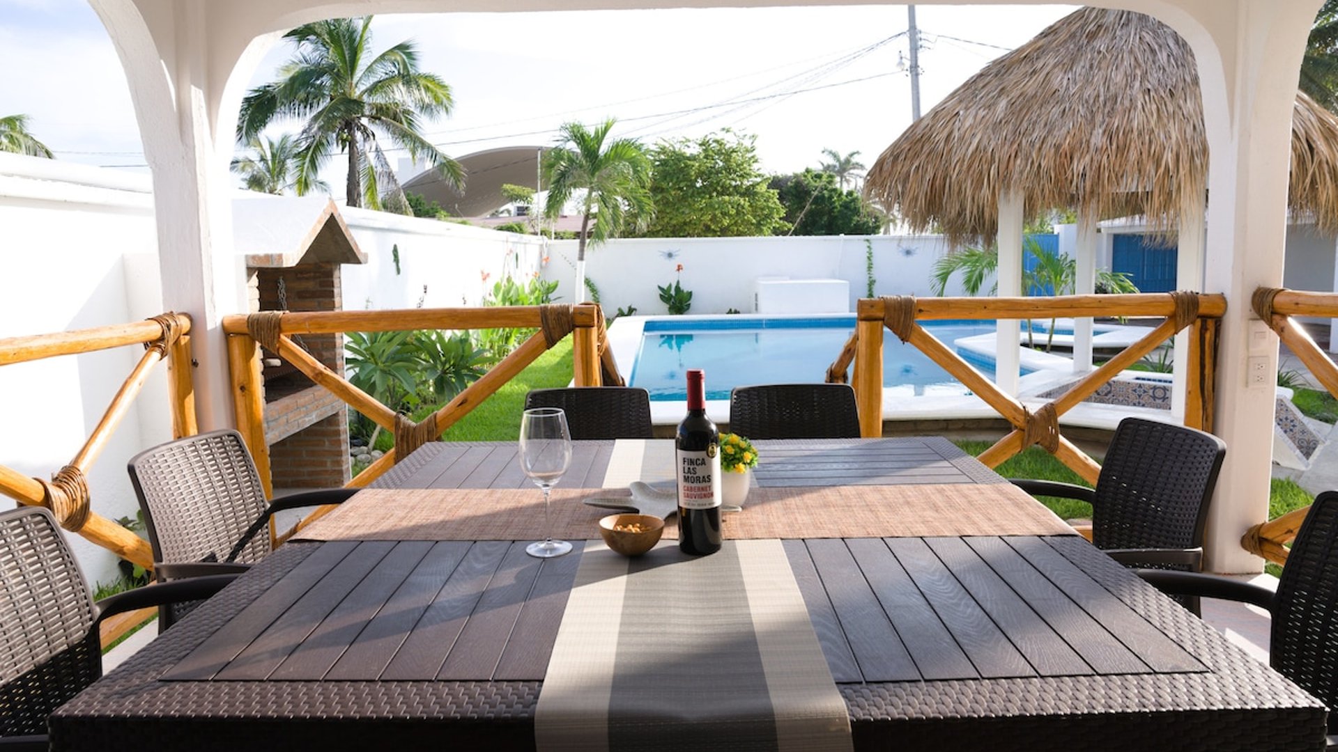 Comedor exterior techado con mesa de seis lugares, botella de vino y copa; al fondo alberca, palapa y palmeras en jardín soleado
