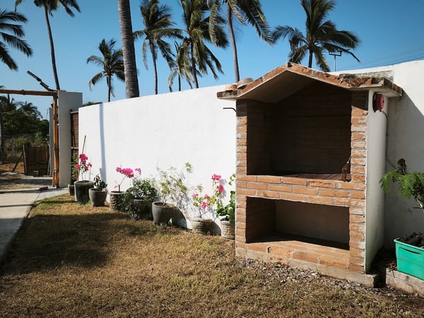 Asador de ladrillo en jardín exterior con buganvilias y palmeras al fondo bajo cielo azul